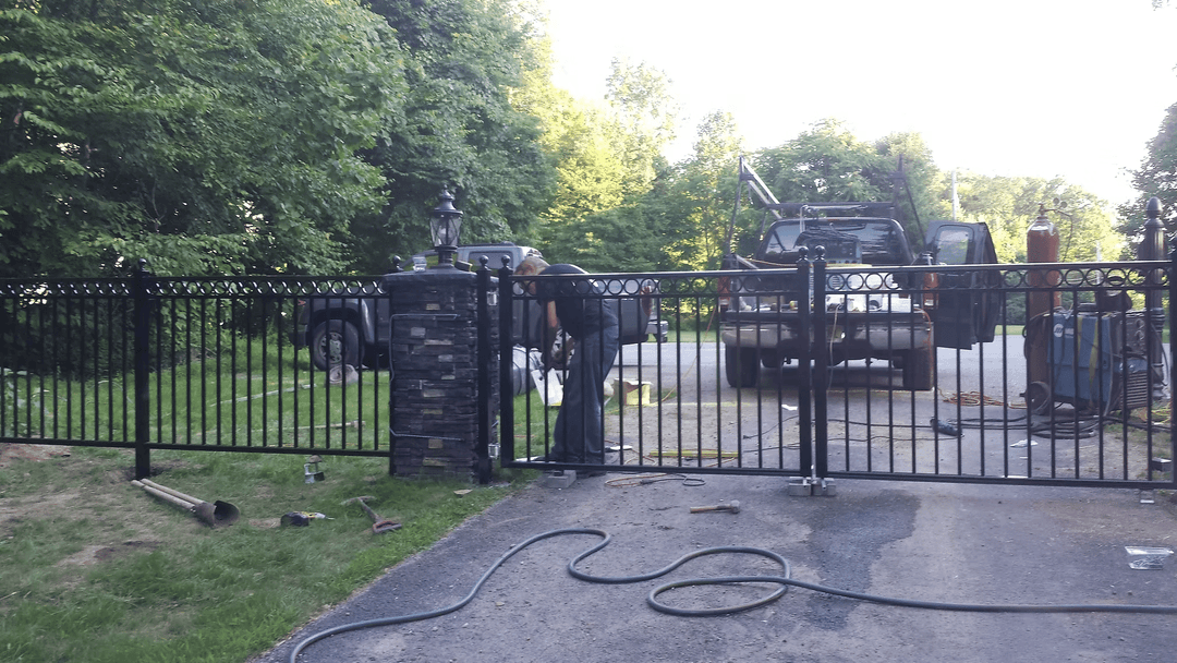 Worker installing a black metal gate in a residential driveway, with a truck nearby.