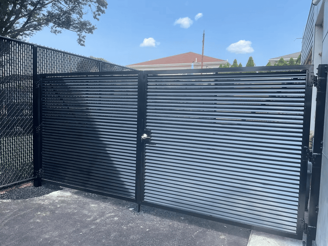 Metal slat gate in a sunny outdoor setting, with a blue sky and residential building in background.