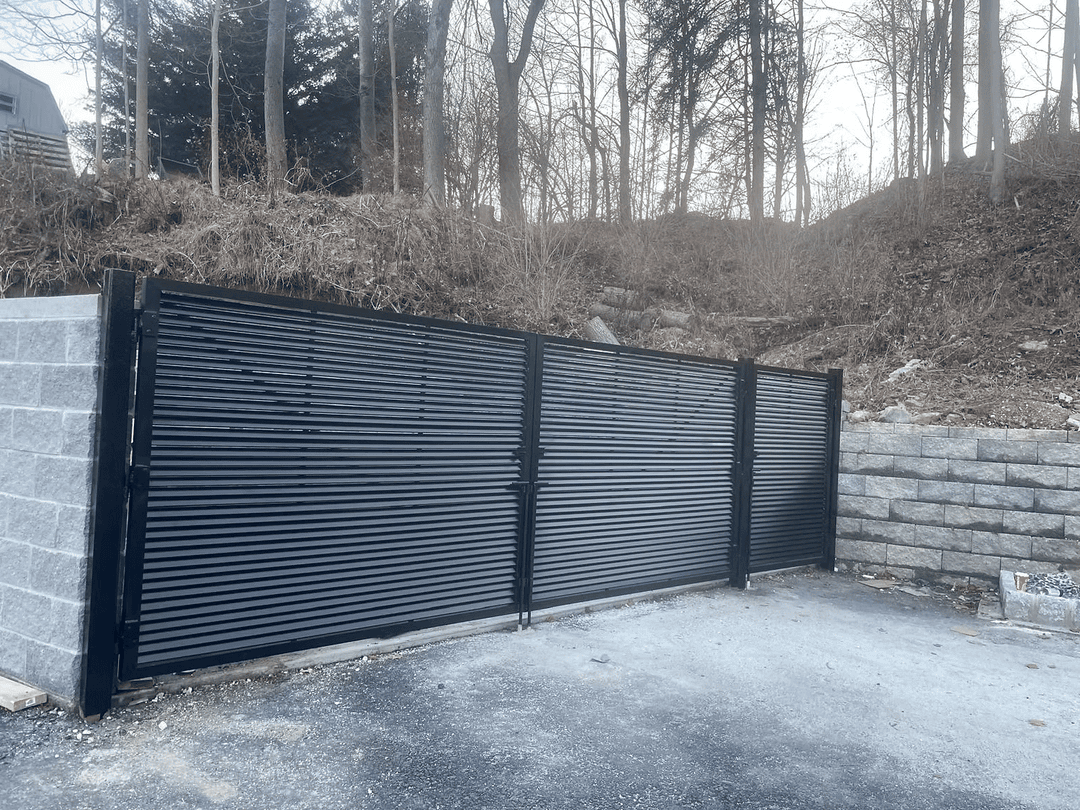 Black horizontal slat gate installed on a gravel driveway near a wooded area.
