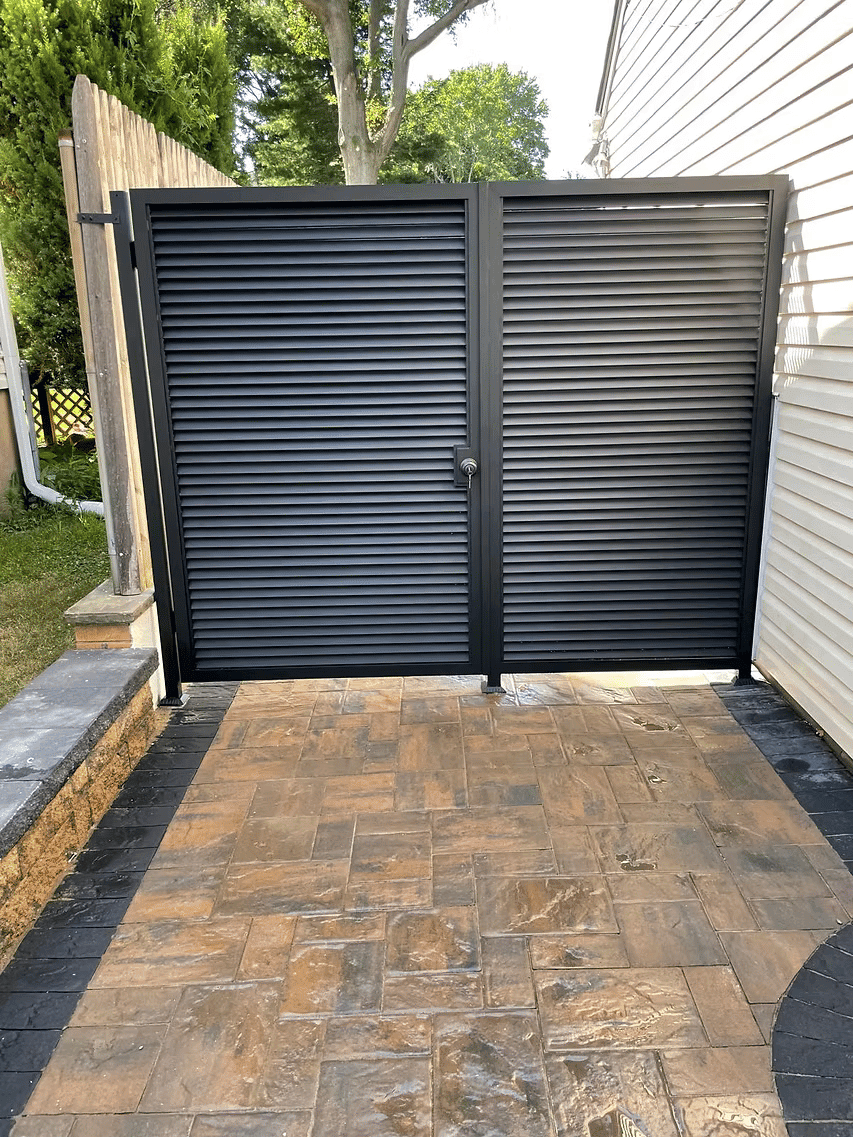 Black slatted gate on a paver patio, leading to a garden area beside a house.