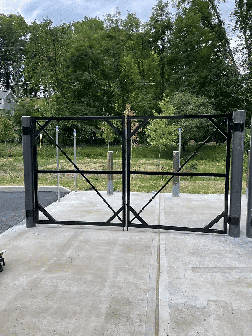 Black metal gate on a concrete path, surrounded by greenery and trees in the background.