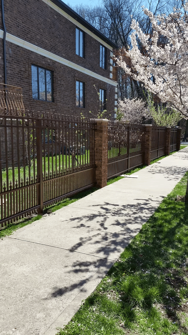 Decorative metal fence along a sidewalk beside an apartment building and blooming trees.