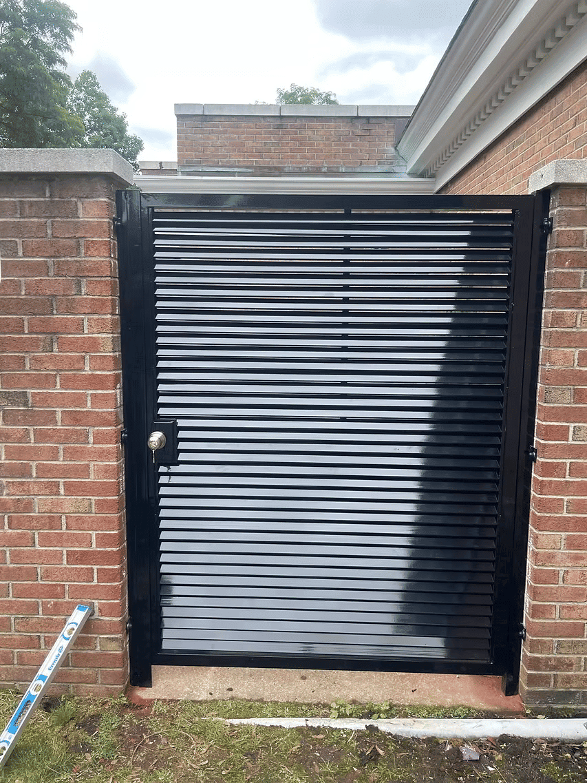 Black metal slatted gate with lock, surrounded by brick wall and greenery.