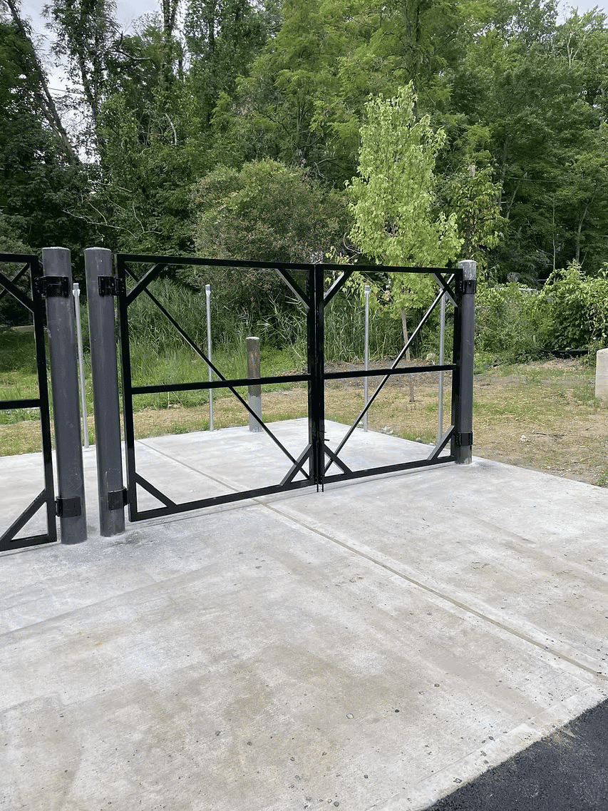 Black metal gate opened on a concrete pathway, surrounded by green vegetation.