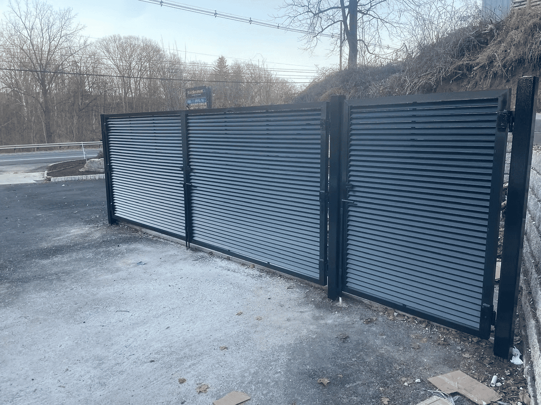 Steel sliding gate with horizontal slats, positioned on a gravel surface near a roadway.
