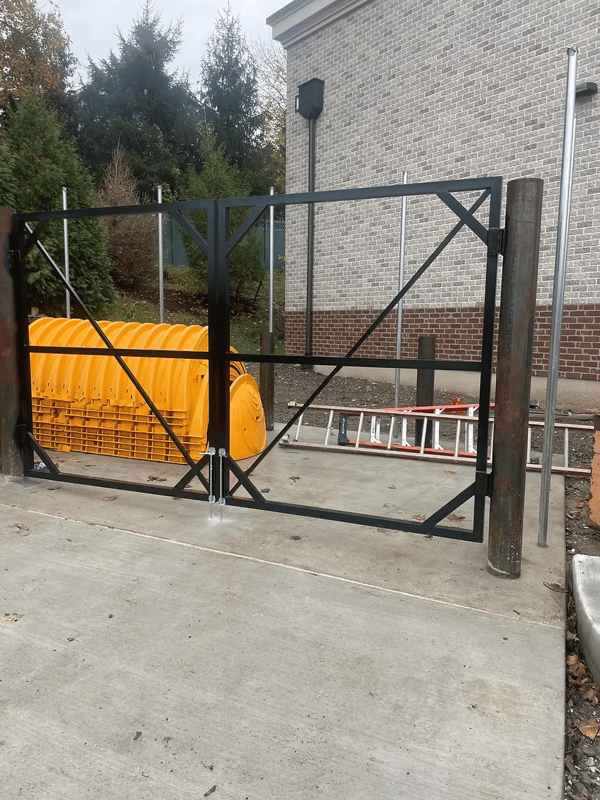 Black metal gate with wooden posts near construction equipment and a ladder on pavement.