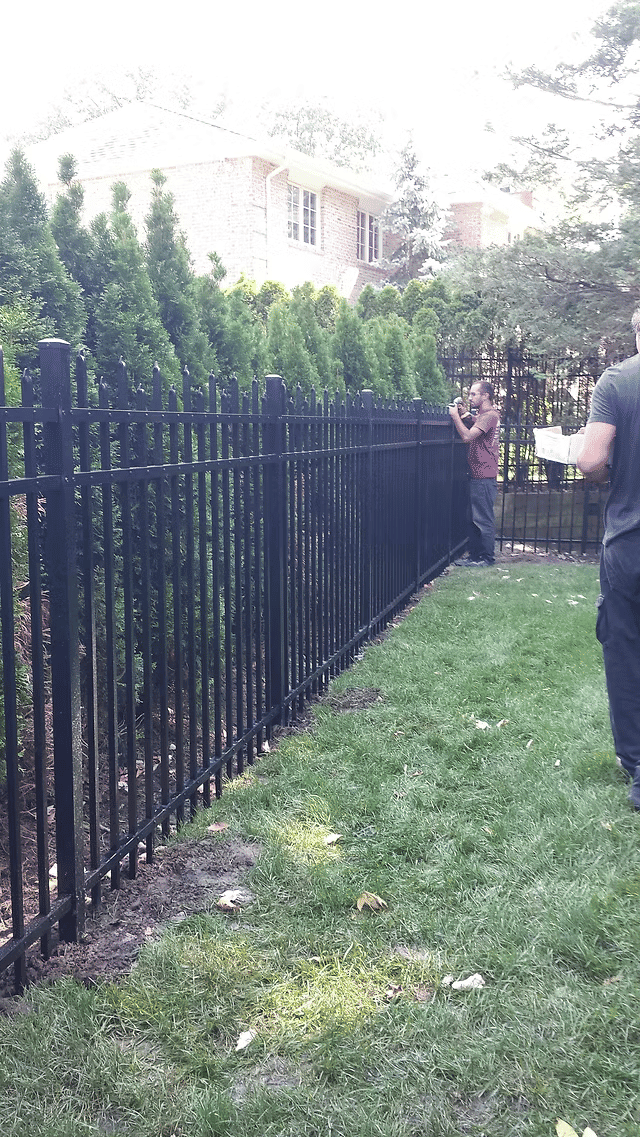 Person installing black metal fence alongside green shrubs in residential yard.