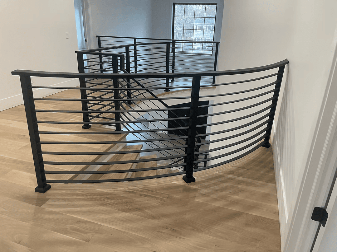 Modern black metal railing curving down stairs in a bright, open space with wooden flooring.