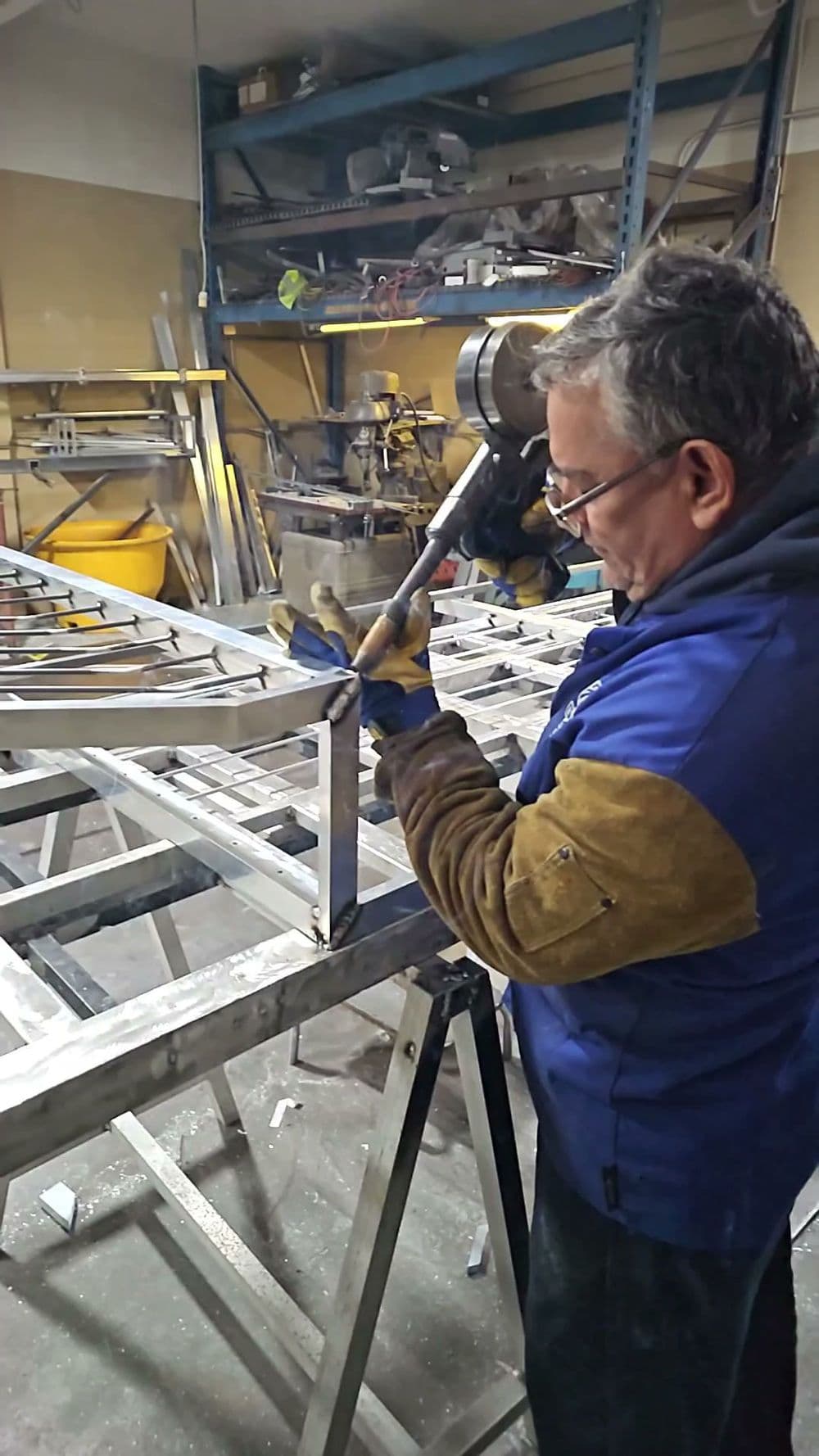 Man welding metal frame in workshop, wearing gloves and protective eyewear.