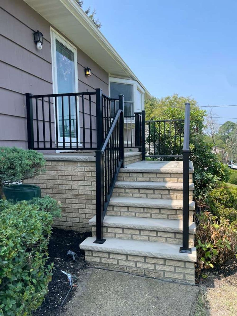 Metal railing on brick steps leading to a house entrance, surrounded by landscaping.