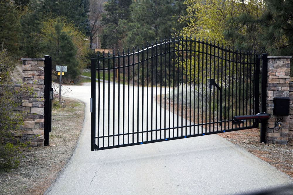 Black wrought iron gate opening on a gravel driveway surrounded by trees and greenery.