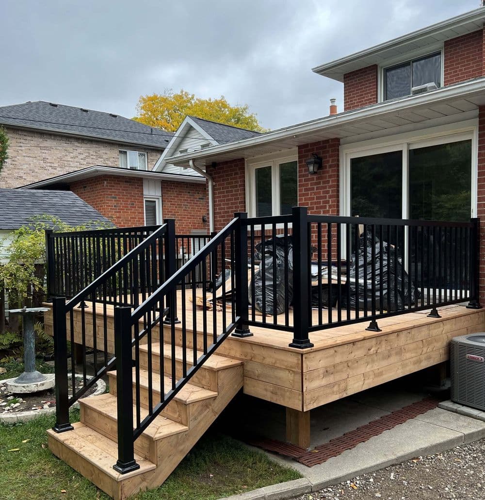 Black railings surround a wooden deck with stairs, adjacent to a brick house and cloudy sky.