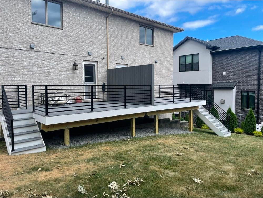 Modern backyard deck with black railing and staircase, surrounded by green lawn and houses.