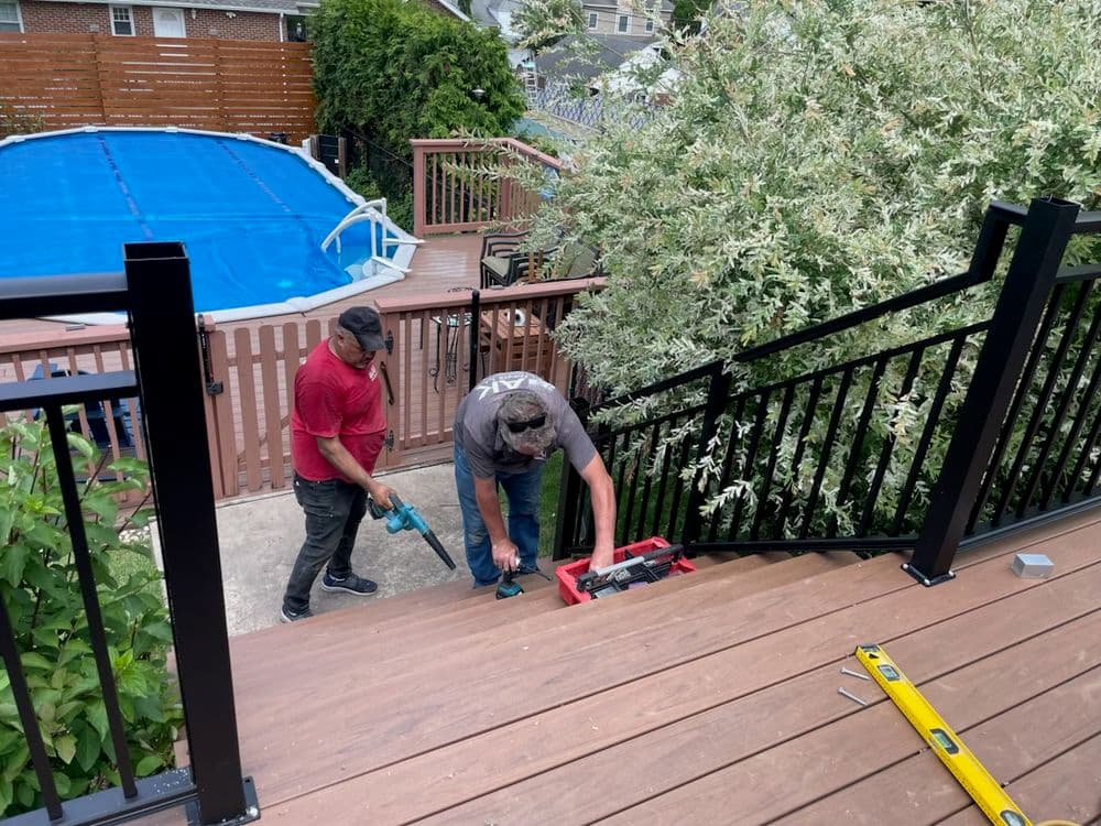 Two men working on a staircase railing installation near a pool in a backyard setting.