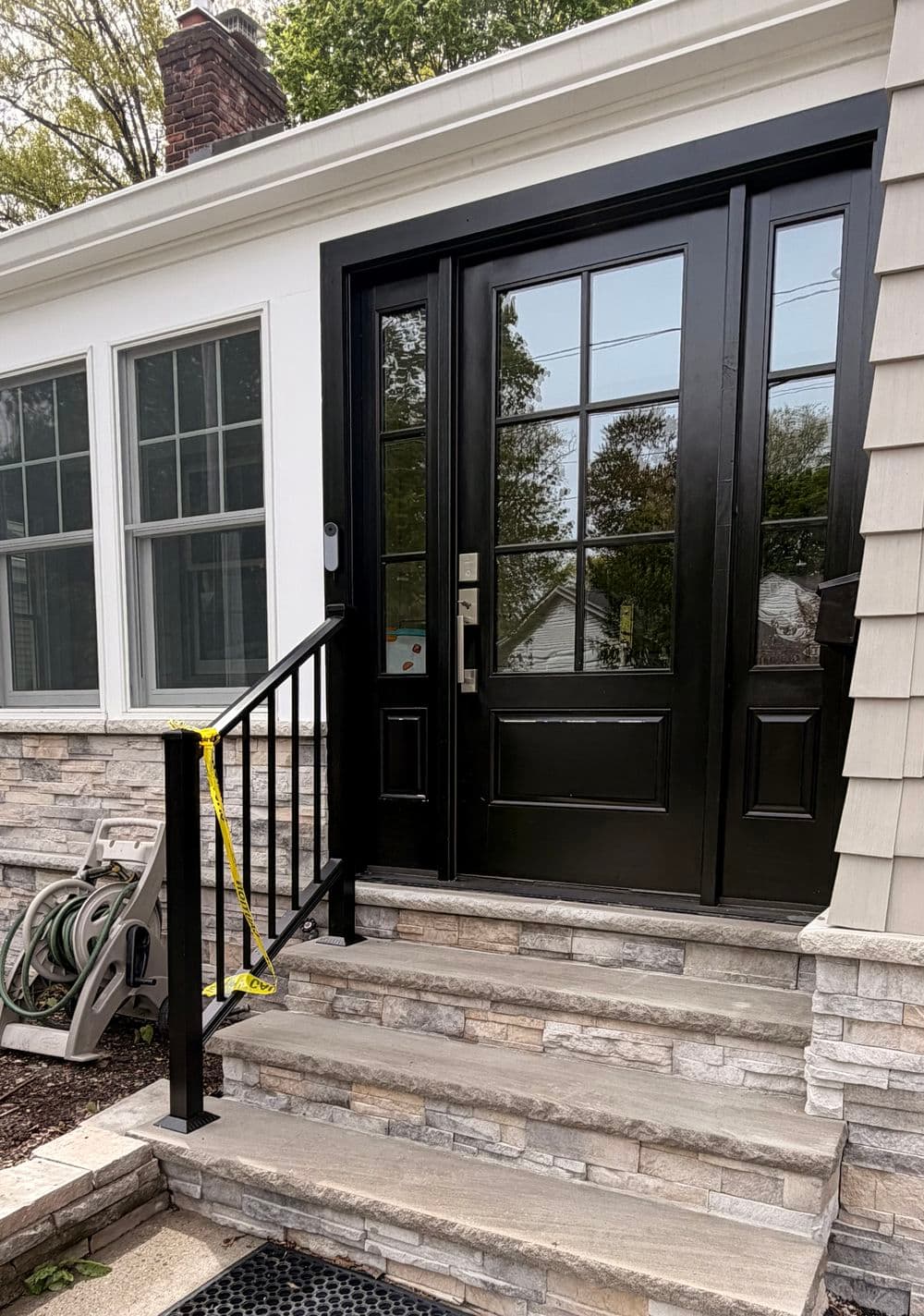 Modern black front door with large windows, stone steps, and railing in a residential setting.