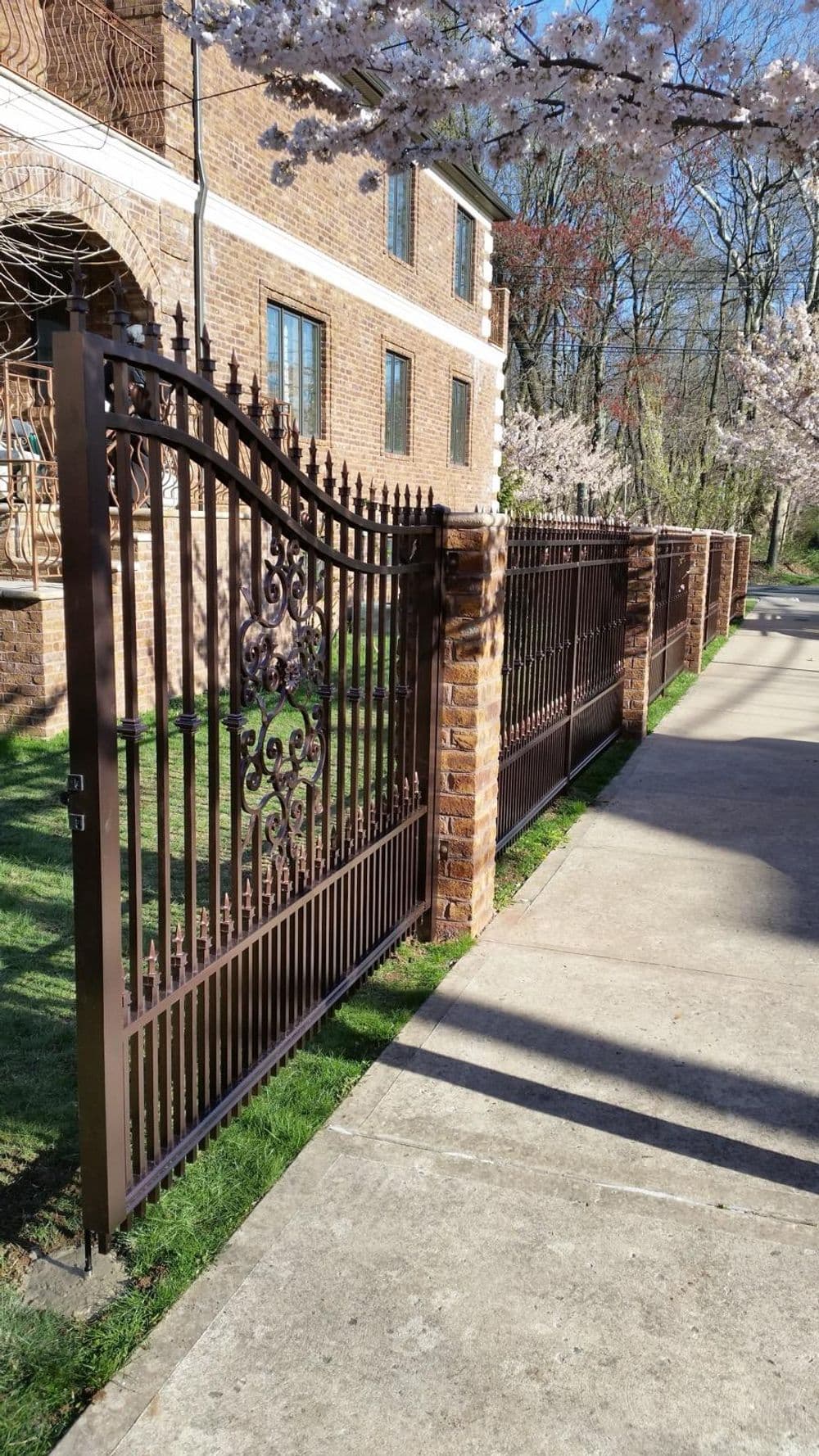 Elegant wrought iron fence with brick pillars, surrounded by flowering trees and a sidewalk.