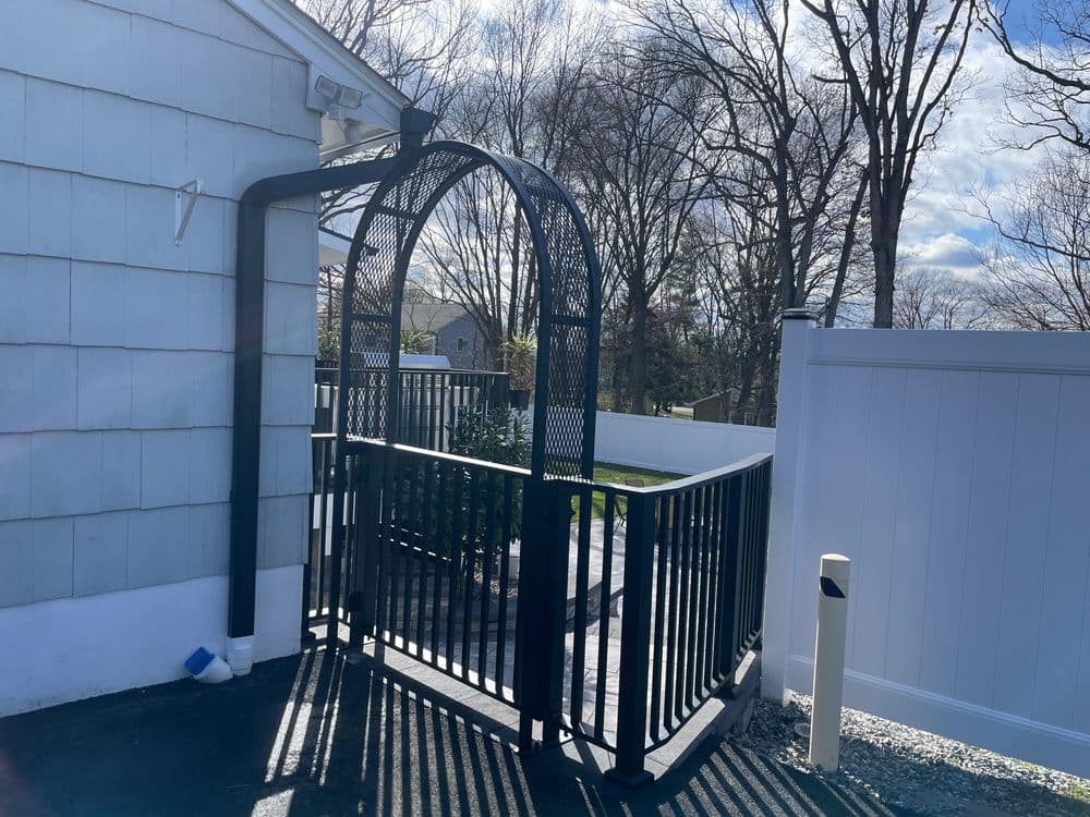 Black metal archway gate leading to a landscaped area with white fencing and clear blue sky.