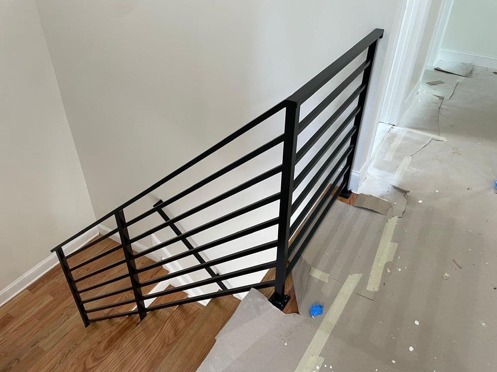 Modern black metal staircase railing installation on wooden floor with unfinished drywall.