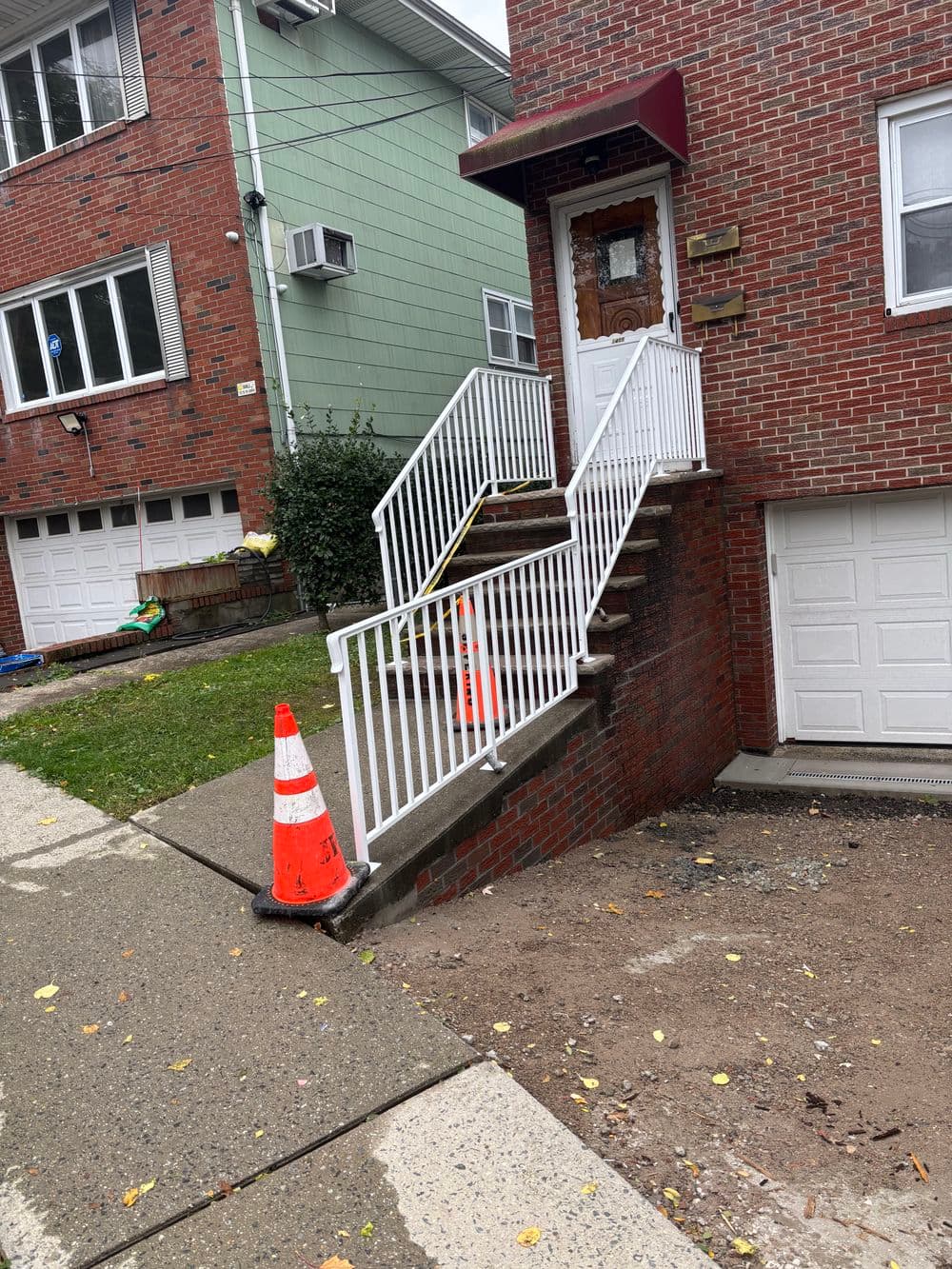 Staircase with white railing leading to a brick house, orange cone on the sidewalk.