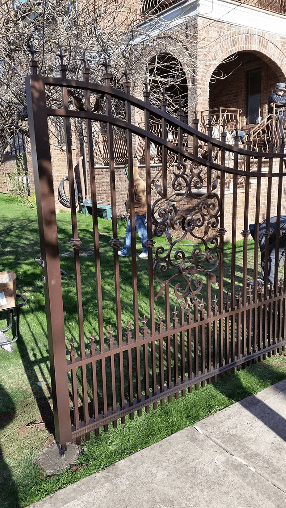 Wrought iron gate with decorative design, leading to a landscaped yard and brick building.