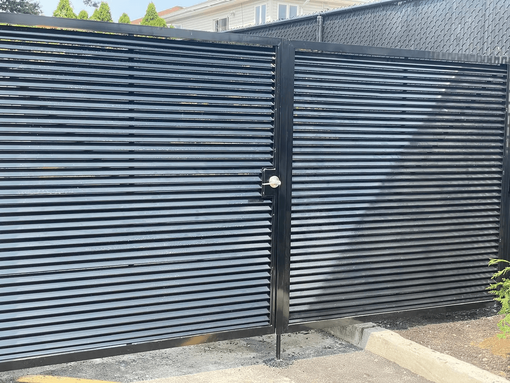 Black metal gate with horizontal slats, leading to an enclosed area, surrounded by greenery.