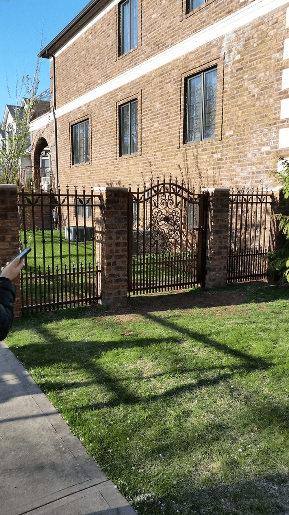 Iron gate beside a brick building and manicured lawn with a person using a smartphone.