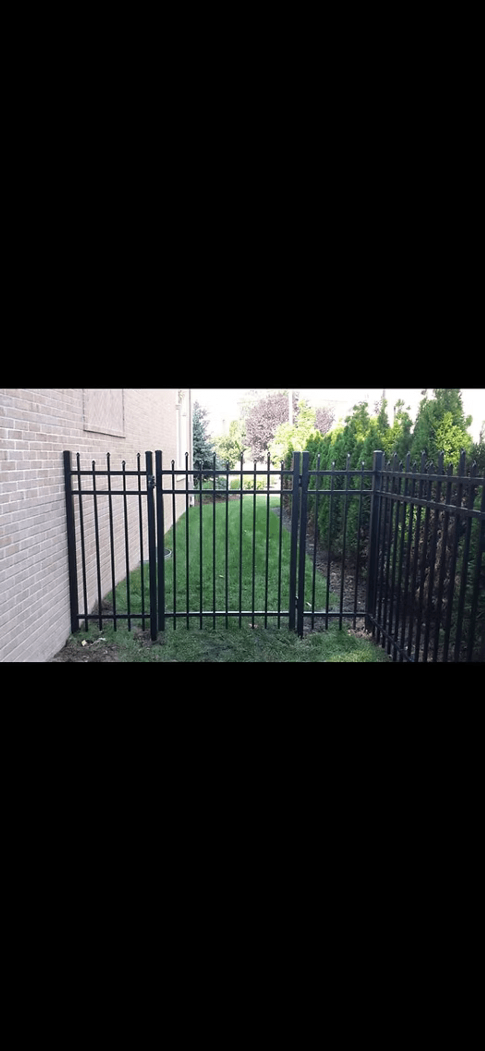 Black metal fence gate leading to a green lawn bordered by shrubs and trees.
