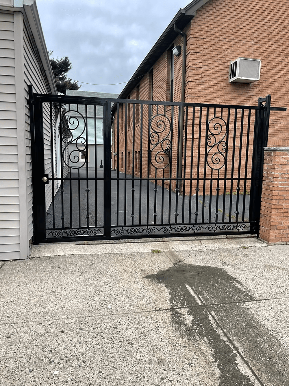 Black wrought iron gate with decorative patterns, leading to a brick building courtyard.