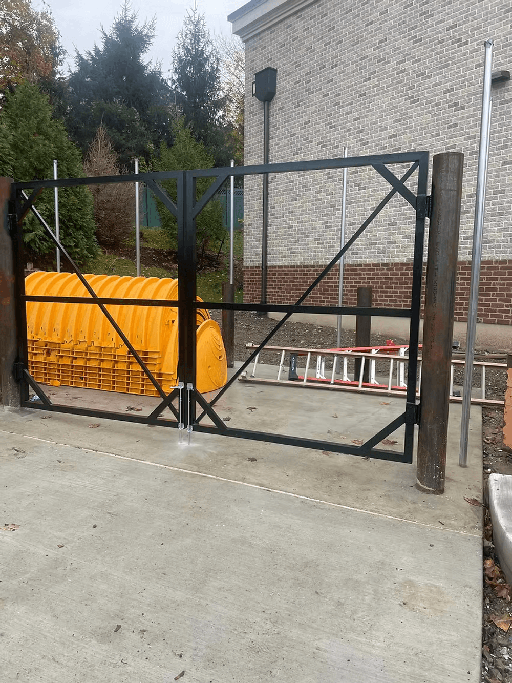 Black metal gate with orange construction tubes and ladder against a brick building.