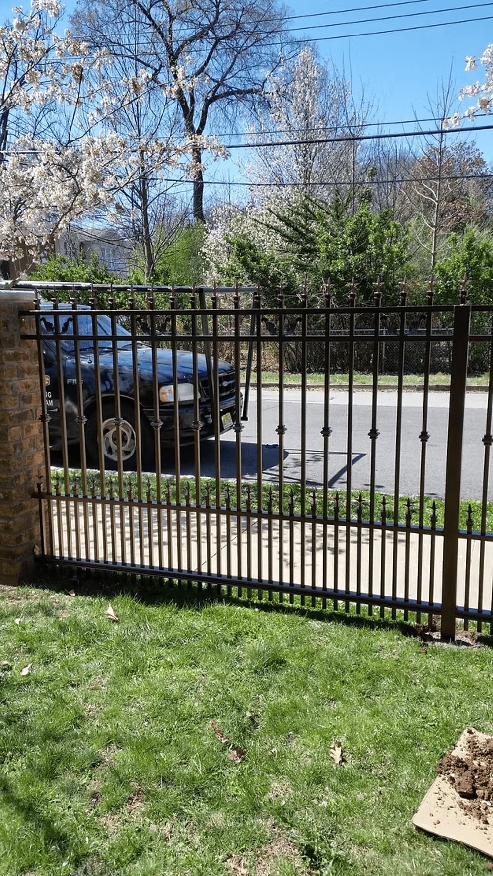 Wrought iron fence with blossoming trees and a parked car on a sunny day.