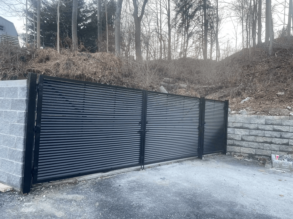 Modern black metal sliding gate at a gravel entrance, surrounded by trees and rocky terrain.