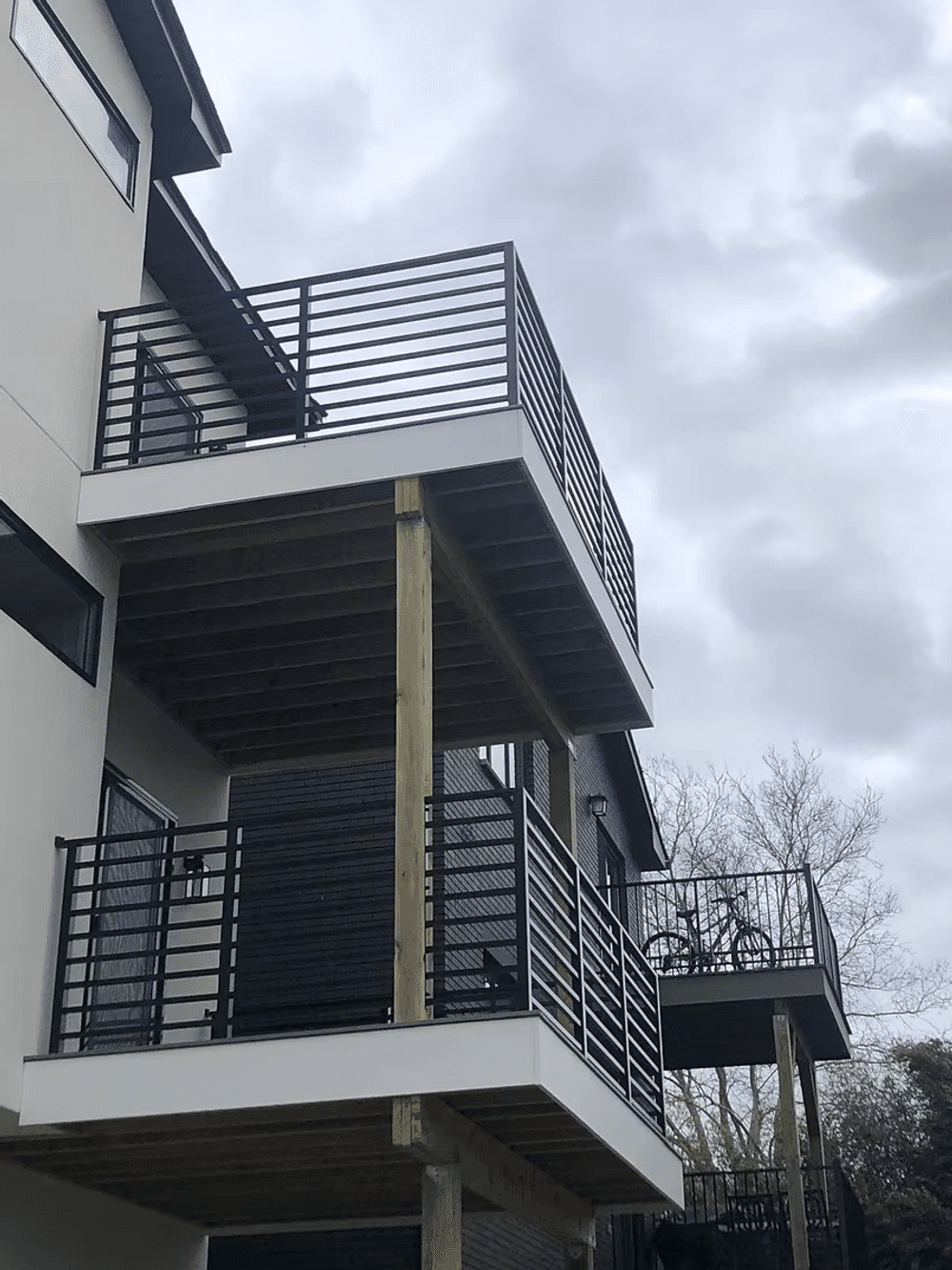 Modern two-story balcony with wooden supports and black metal railings against a cloudy sky.