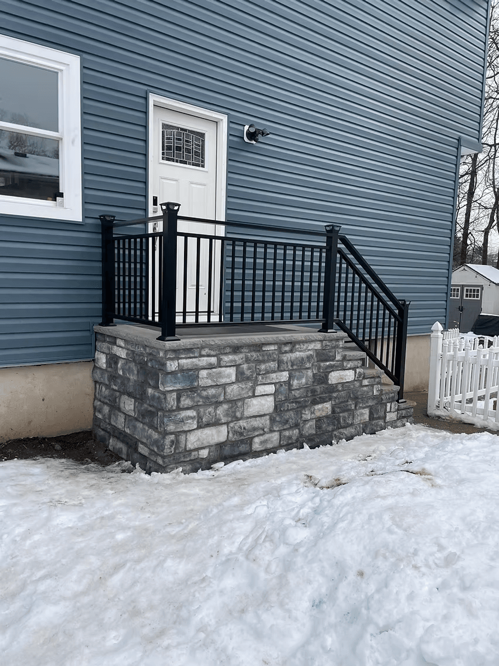 Stone steps with black railing leading to a white door on a blue house, surrounded by snow.