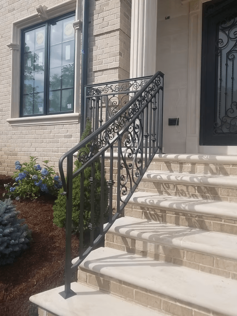 Elegant black wrought iron railing on stone steps beside a brick home, featuring floral landscaping.