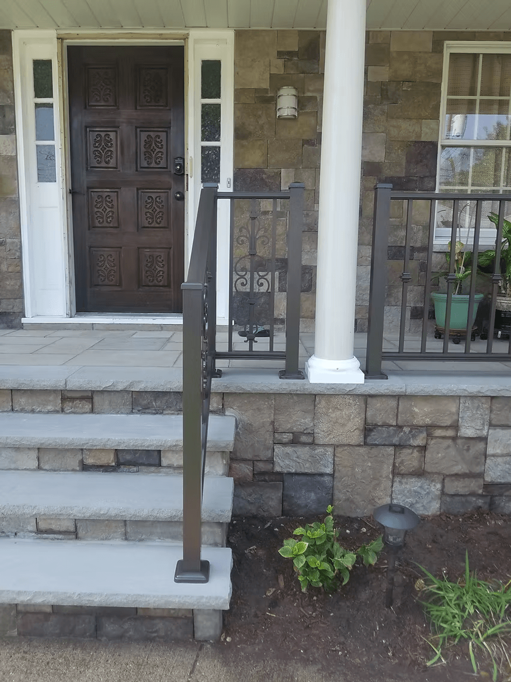 Front porch with stone steps, iron railing, and a wooden door. Potted plants add greenery.