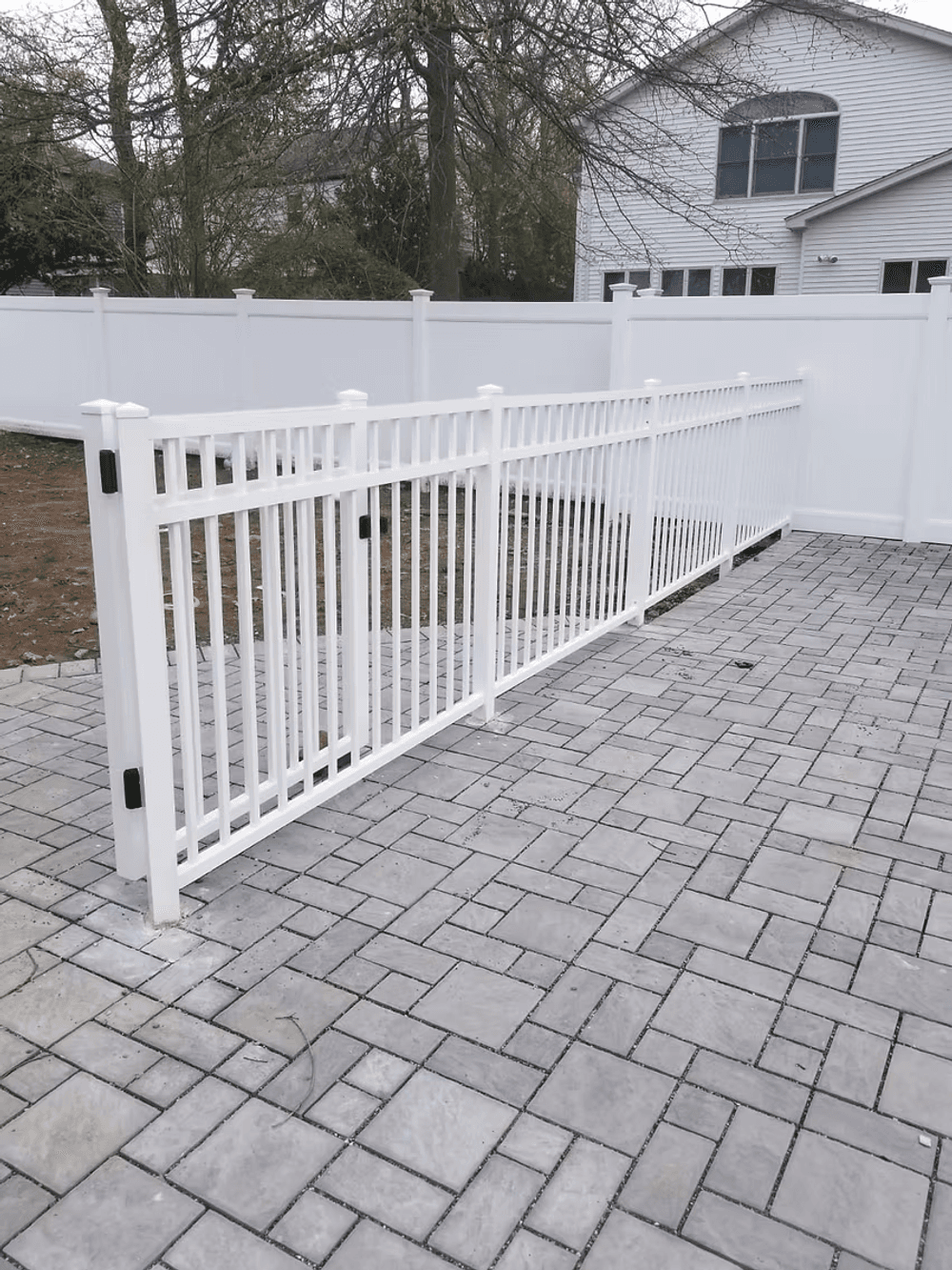 White vinyl fence enclosing a stone patio area with a house and trees in the background.
