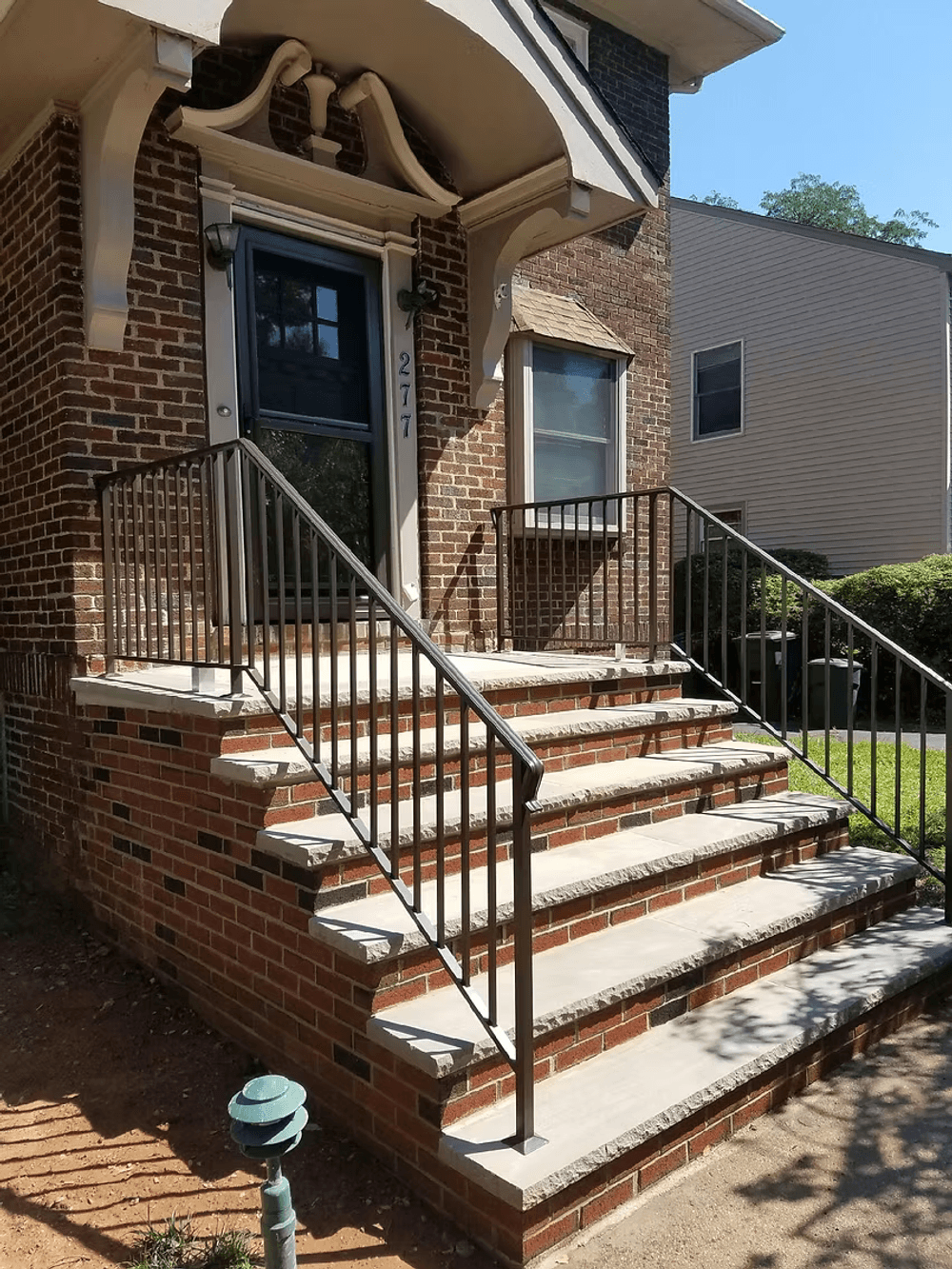 Brick building entrance with steps and metal railing, featuring clear sunlight and greenery.