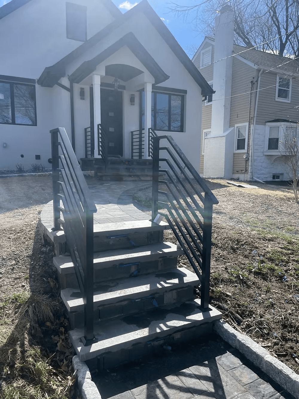 Modern home entrance with stone steps and black railing on a sunny day.