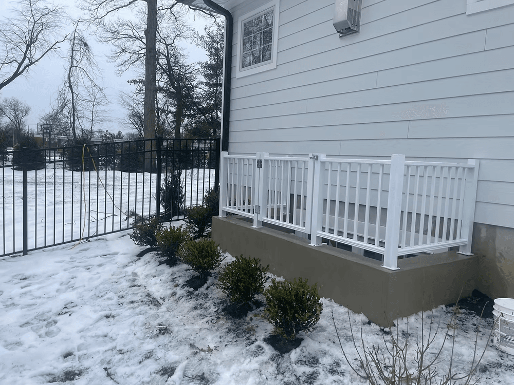 Snowy landscape featuring a white fenced porch and manicured shrubs beside a house.