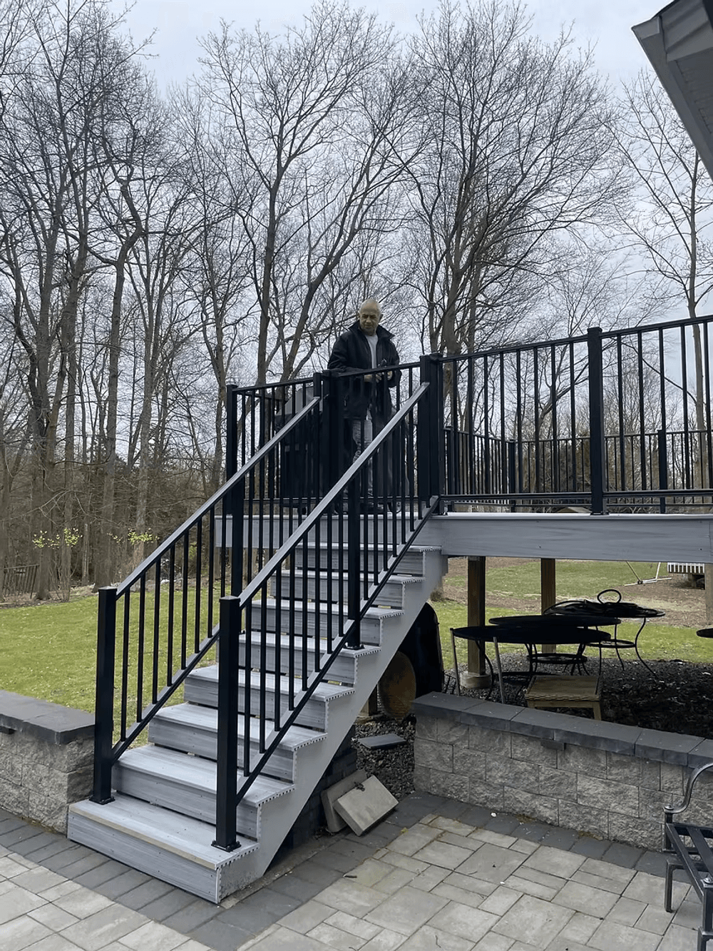 Person standing on a black railing staircase in a backyard with trees in the background.