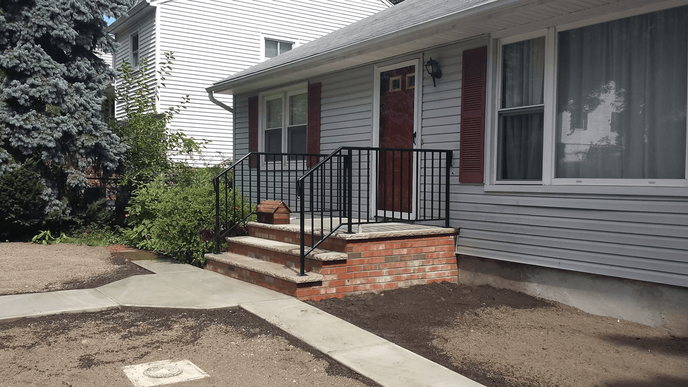 Front entrance of a house featuring brick steps, black railing, and landscaped surroundings.