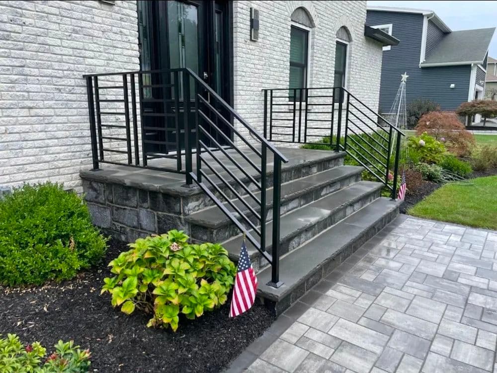 Modern home entrance with stone steps, black railings, and decorative plants. American flags displayed.