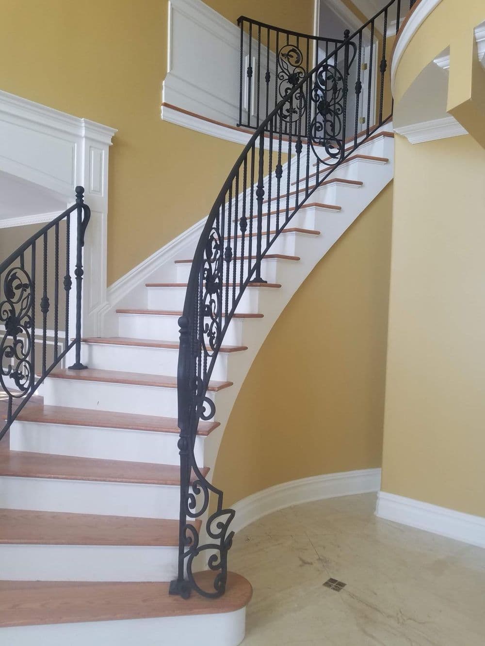 Elegant spiral staircase with wooden steps and ornate black iron railing in a yellow foyer.