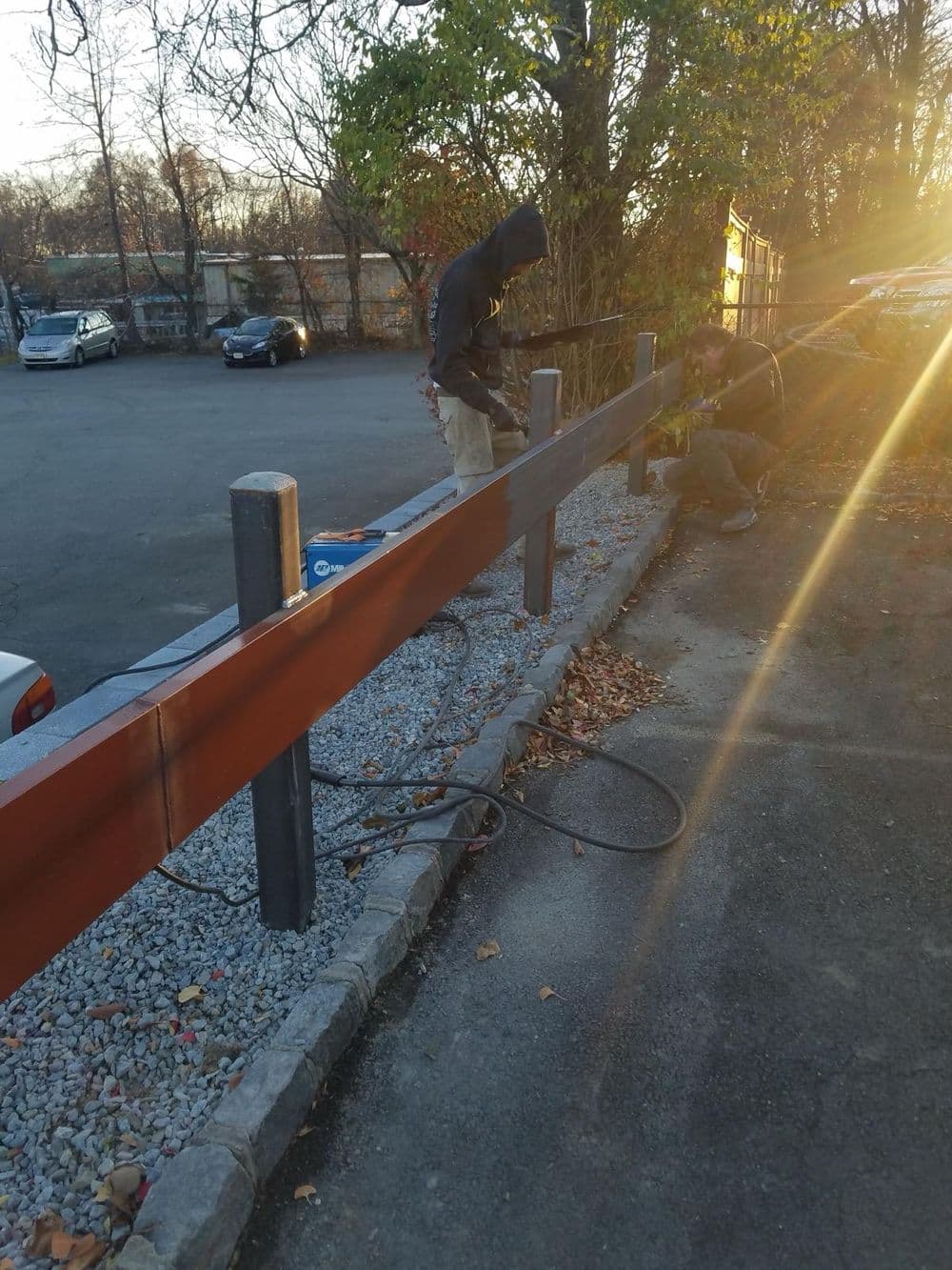 Two workers installing a wooden fence along a gravel pathway in a parking lot.