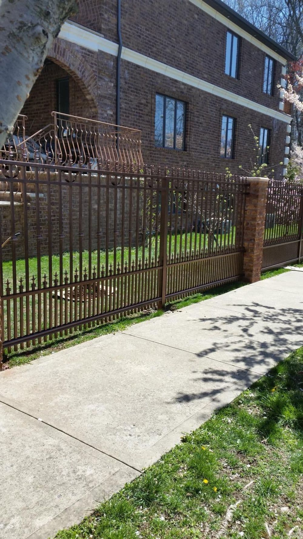Iron fence along a sidewalk in front of a brick house with windows and greenery.