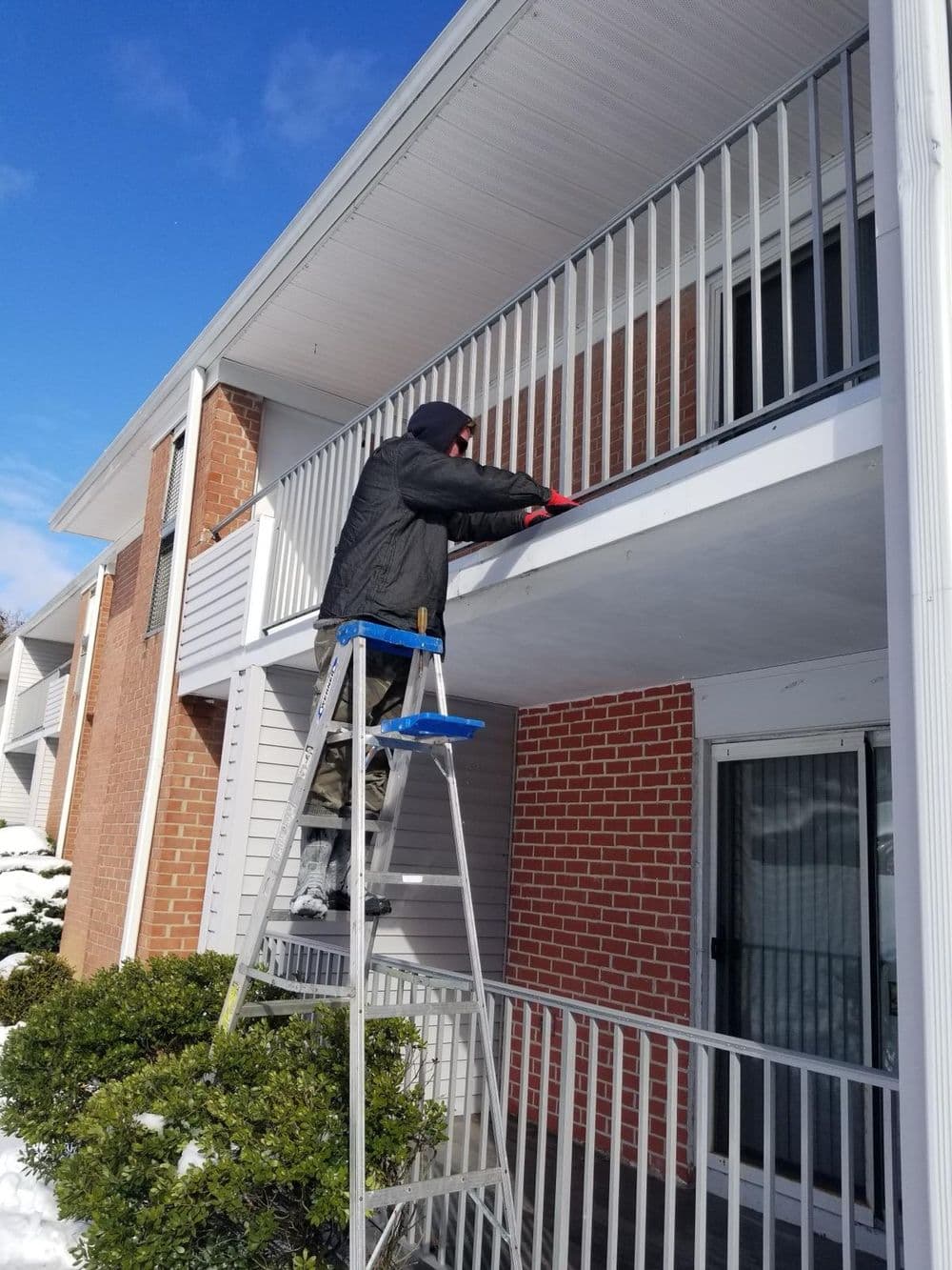 Person using a ladder to maintain a balcony on a snowy day at an apartment building.