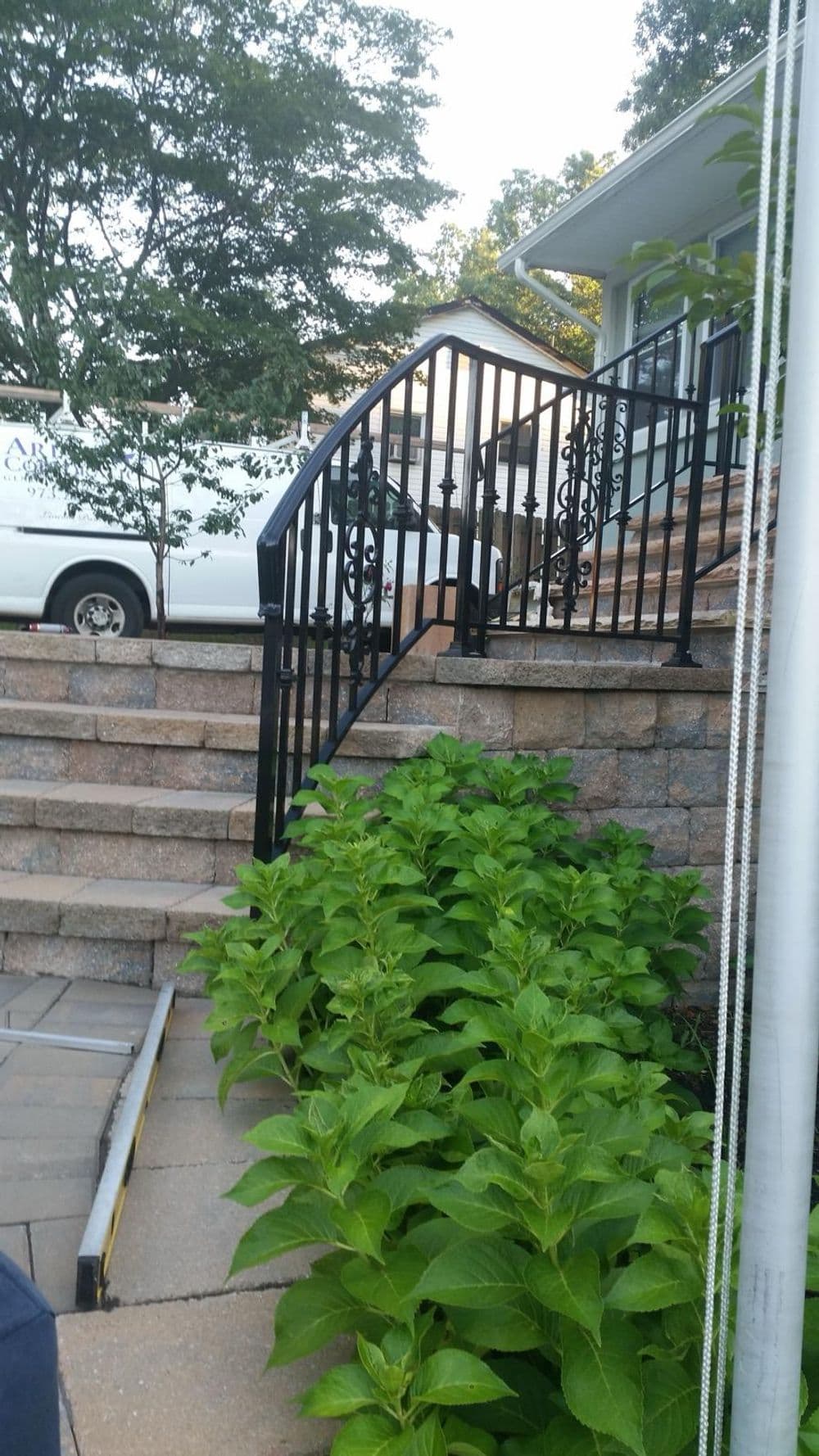 Black metal railing beside stone steps leading to a house, surrounded by lush green plants.