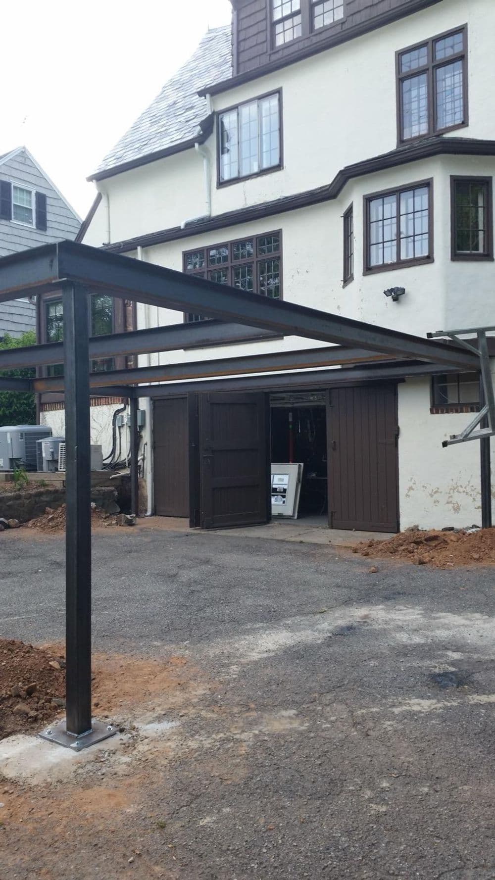 Metal carport structure in front of a large traditional home with brown double doors.
