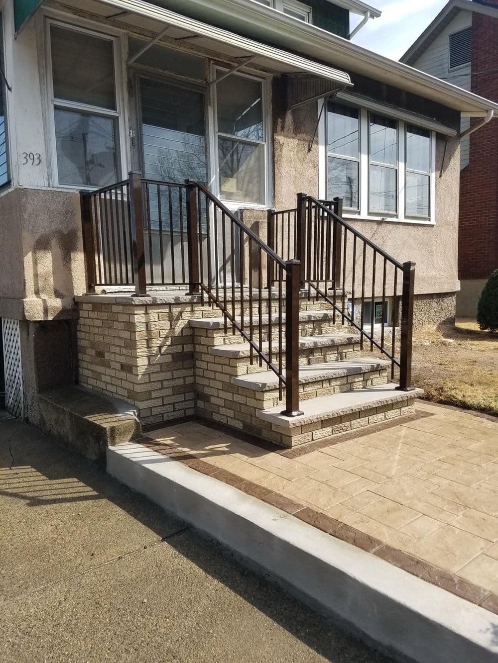 Exterior view of a home entrance with brick steps and black railings, featuring a smooth concrete pathway.