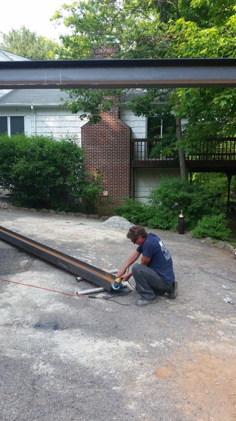 Man working with metal beams outdoors near a house surrounded by greenery.